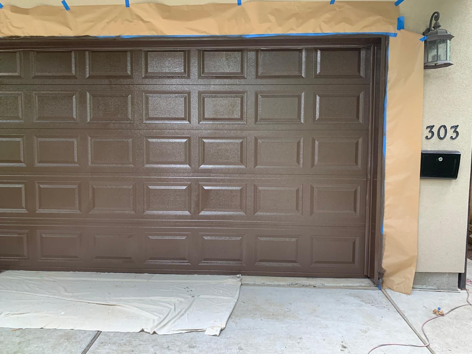 Brown garage door being painted, masked with brown paper and blue tape; house number 303.