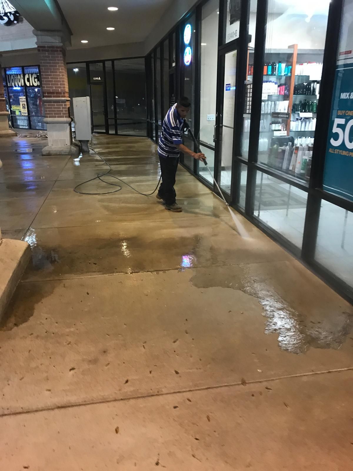 Man pressure washing a concrete sidewalk outside a storefront at night.