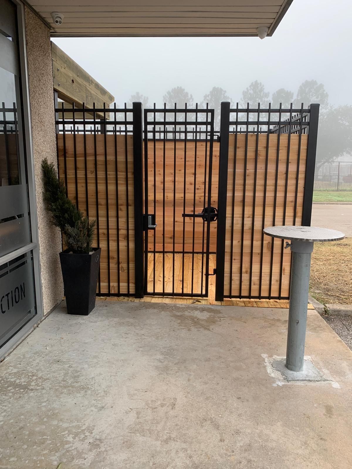 Black metal and wood fence with gate on a concrete patio; a plant in a black pot sits to the left.