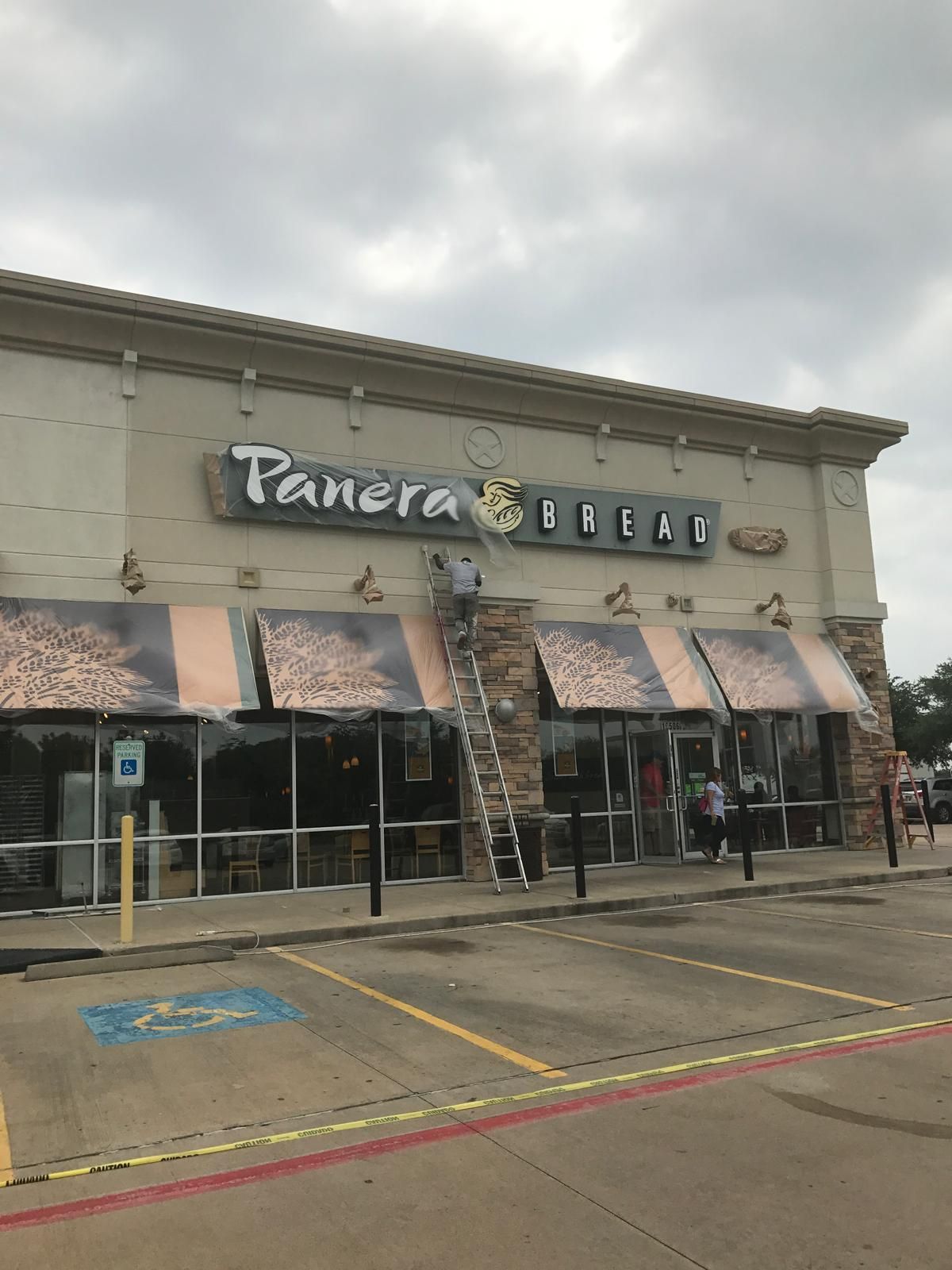 A person on a ladder cleans the Panera Bread sign on a beige building with awnings under a cloudy sky.