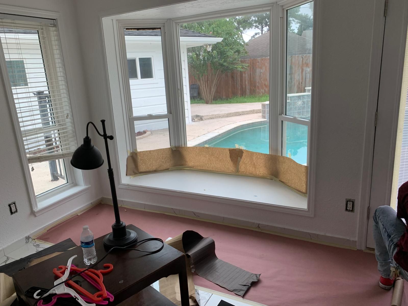 Interior room with bay window overlooking a pool. Pink floor covering, brown table with lamp, and white window frames.