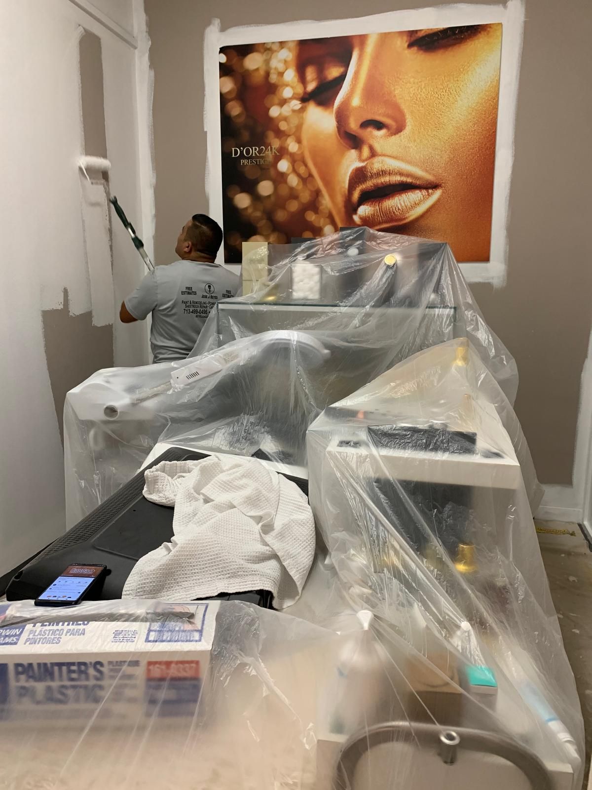 Man painting wall in a room with gold-toned portrait, covered equipment, and plastic sheeting.