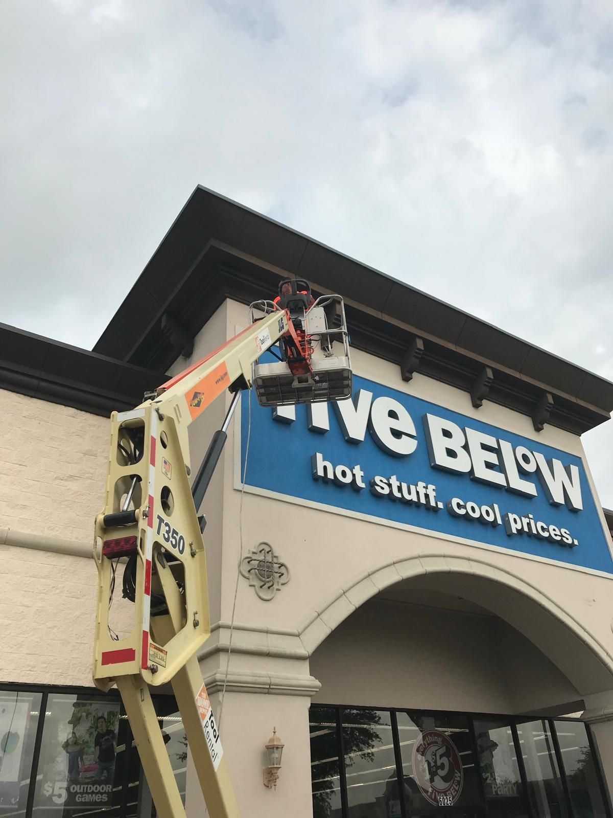 A worker in a lift is working on the Five Below store sign under a cloudy sky.