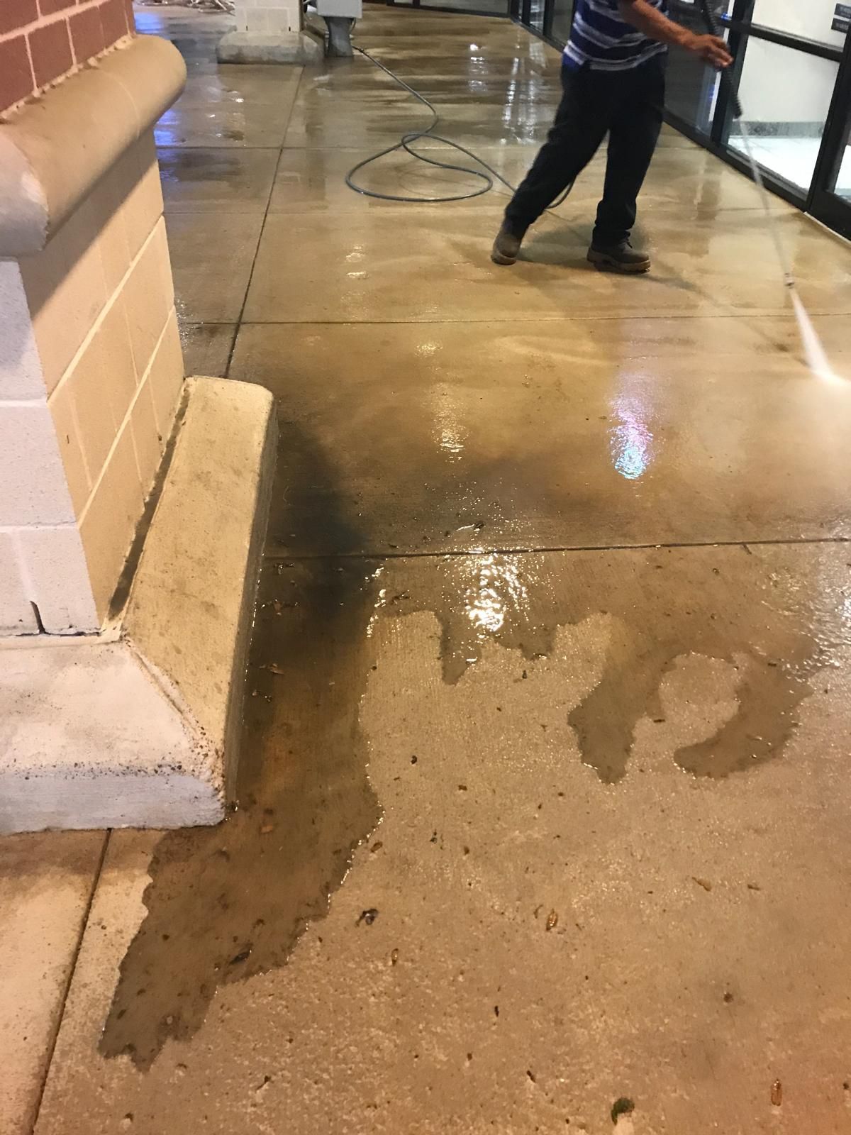 Person power washing a wet concrete sidewalk. Brick building on the left, reflection in the glass.