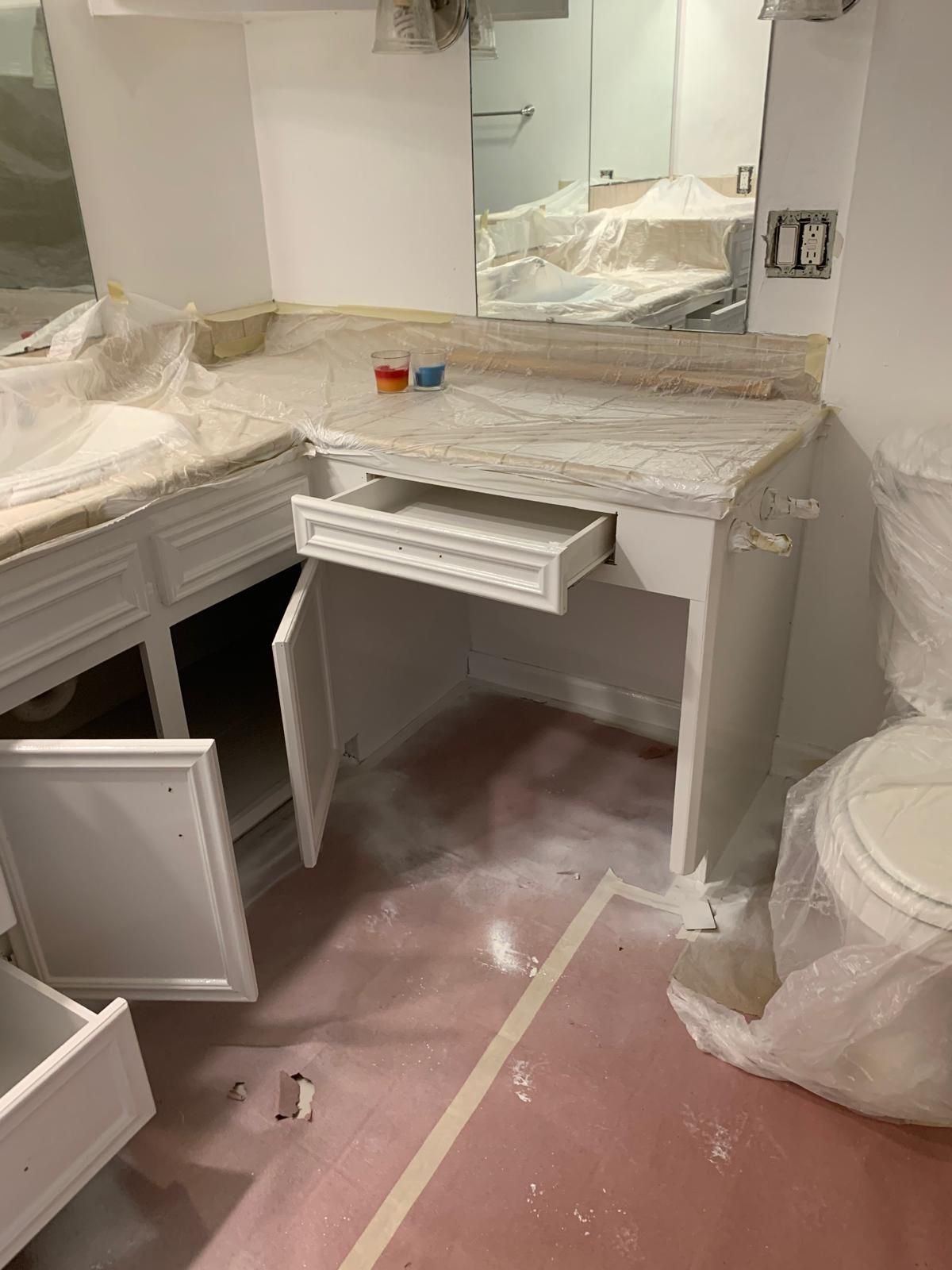 Bathroom vanity under construction; white cabinets, open drawer, protective coverings, pink floor.