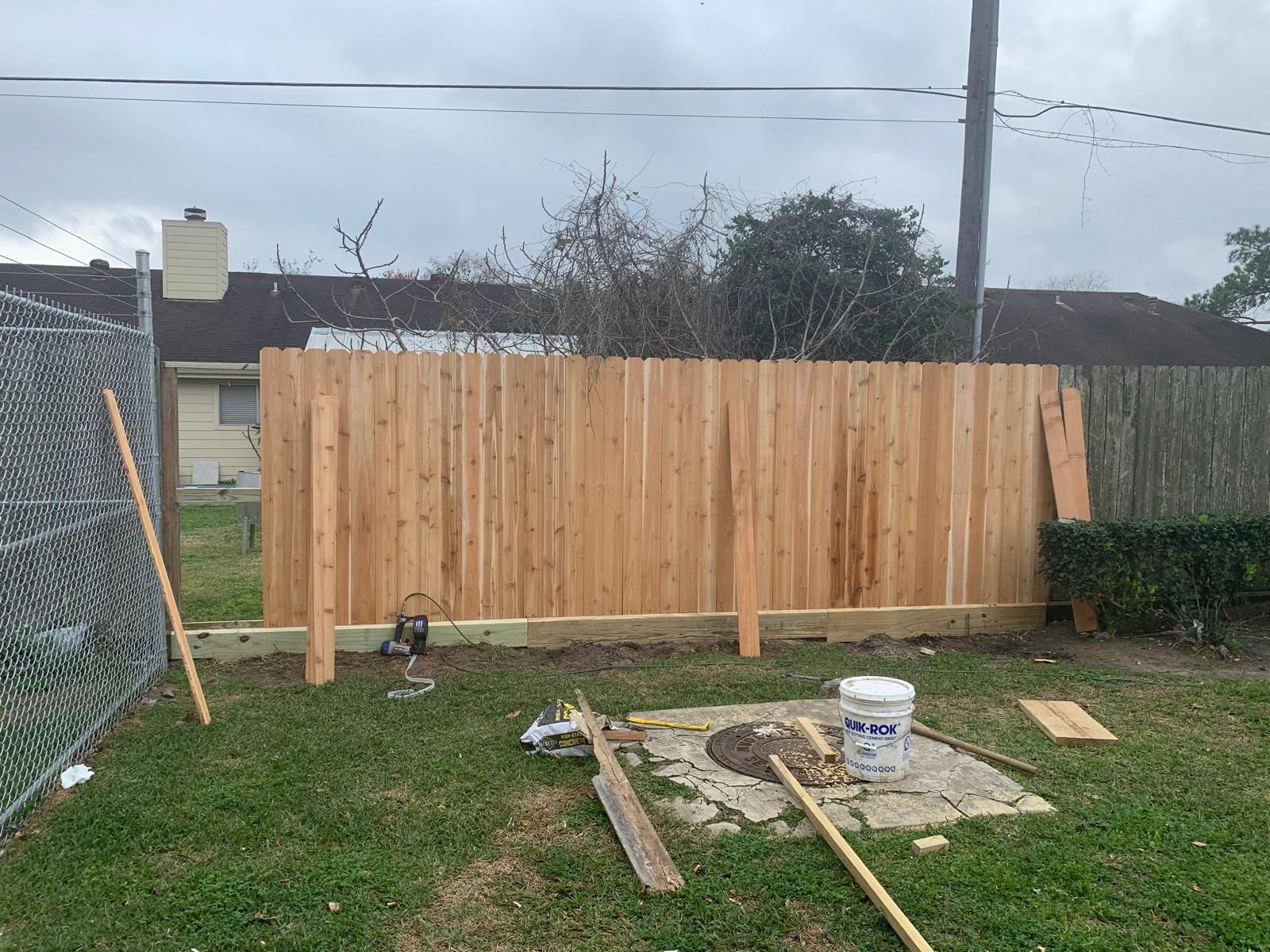 Wooden fence under construction in a grassy backyard. Tools and materials are scattered around. Cloudy sky.