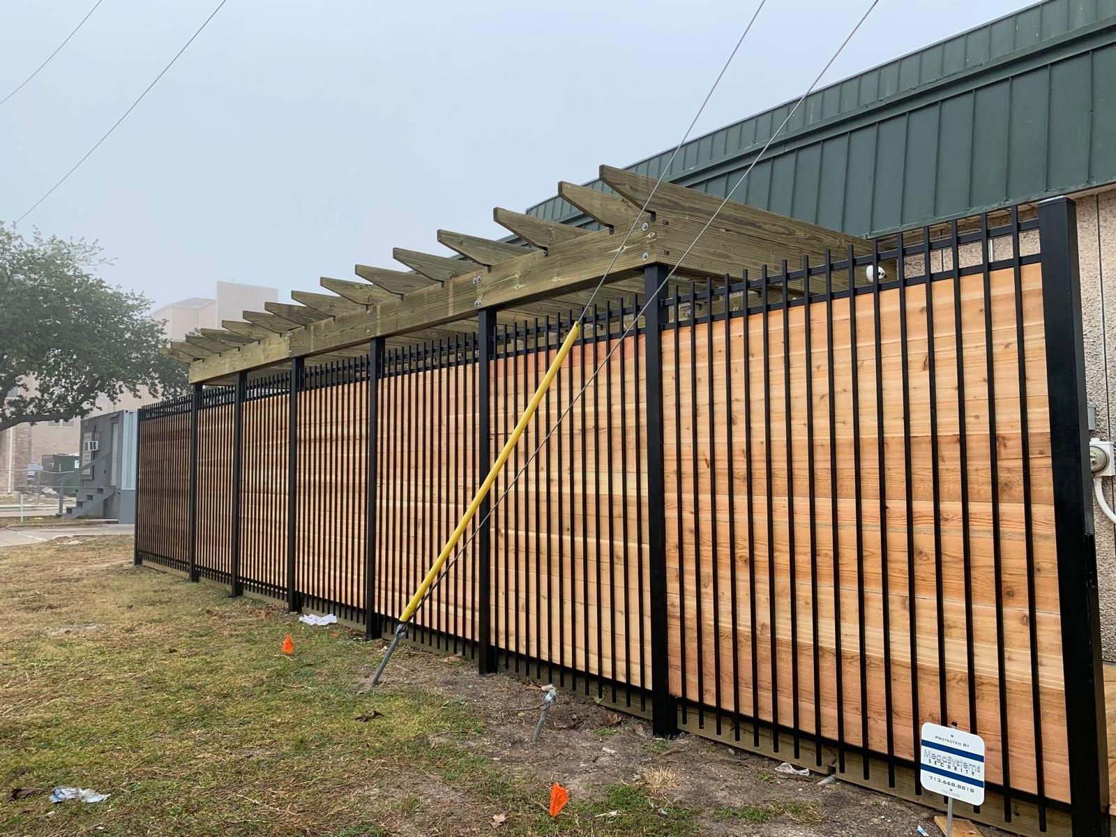 Black metal fence with wooden slats and pergola, alongside a building on a grassy area.