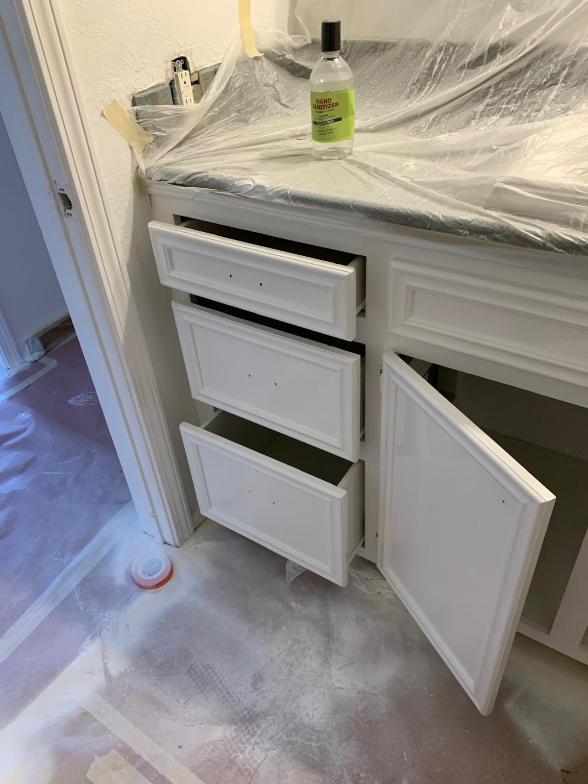 Bathroom vanity with open drawers and door, white paint, countertop covered in plastic.