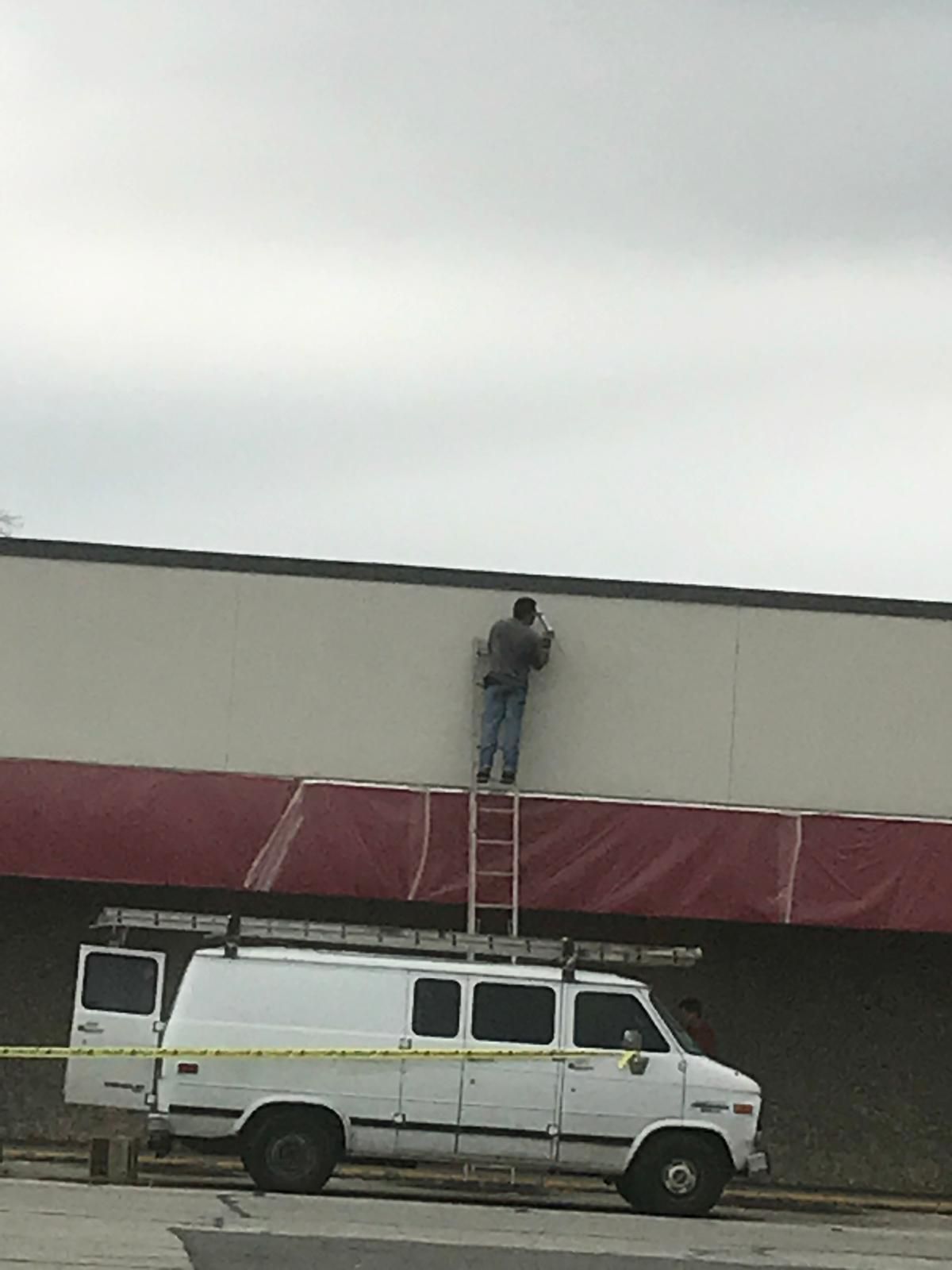 Man on a ladder, working on the exterior of a building, van parked below. Cloudy sky.