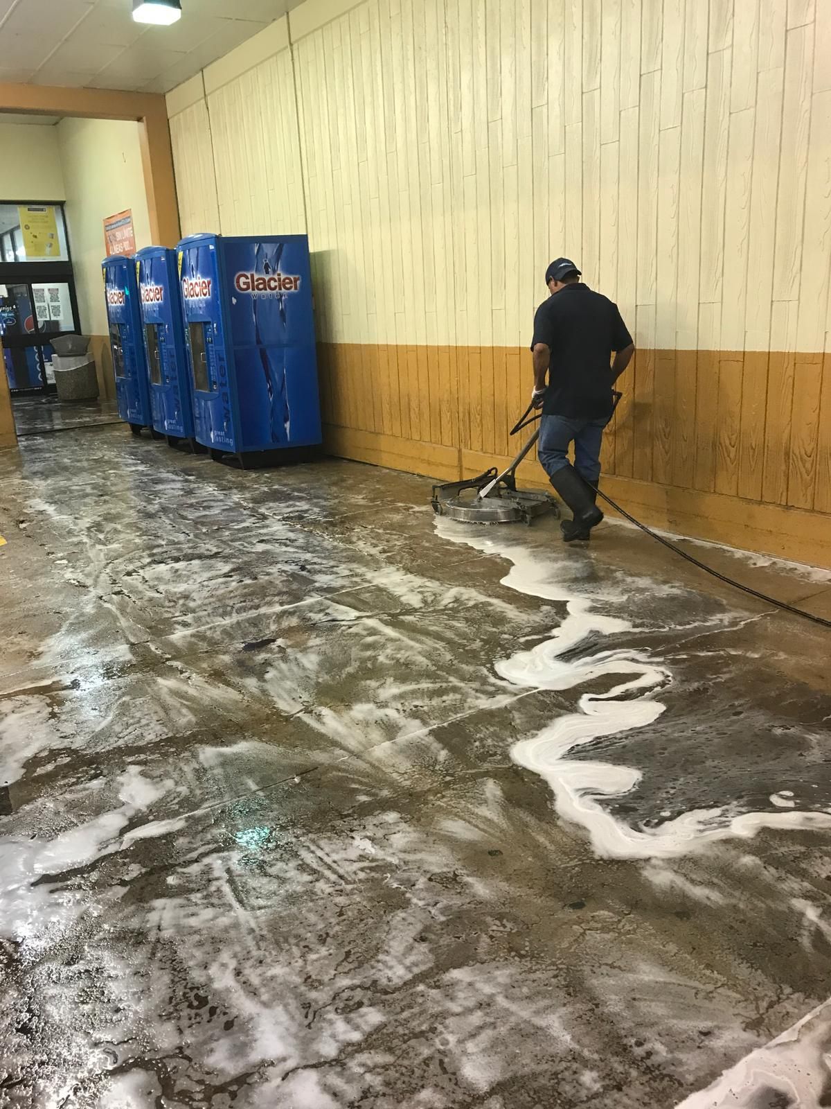 A person pressure washes a floor with soapy water in a store; three blue vending machines line the wall.