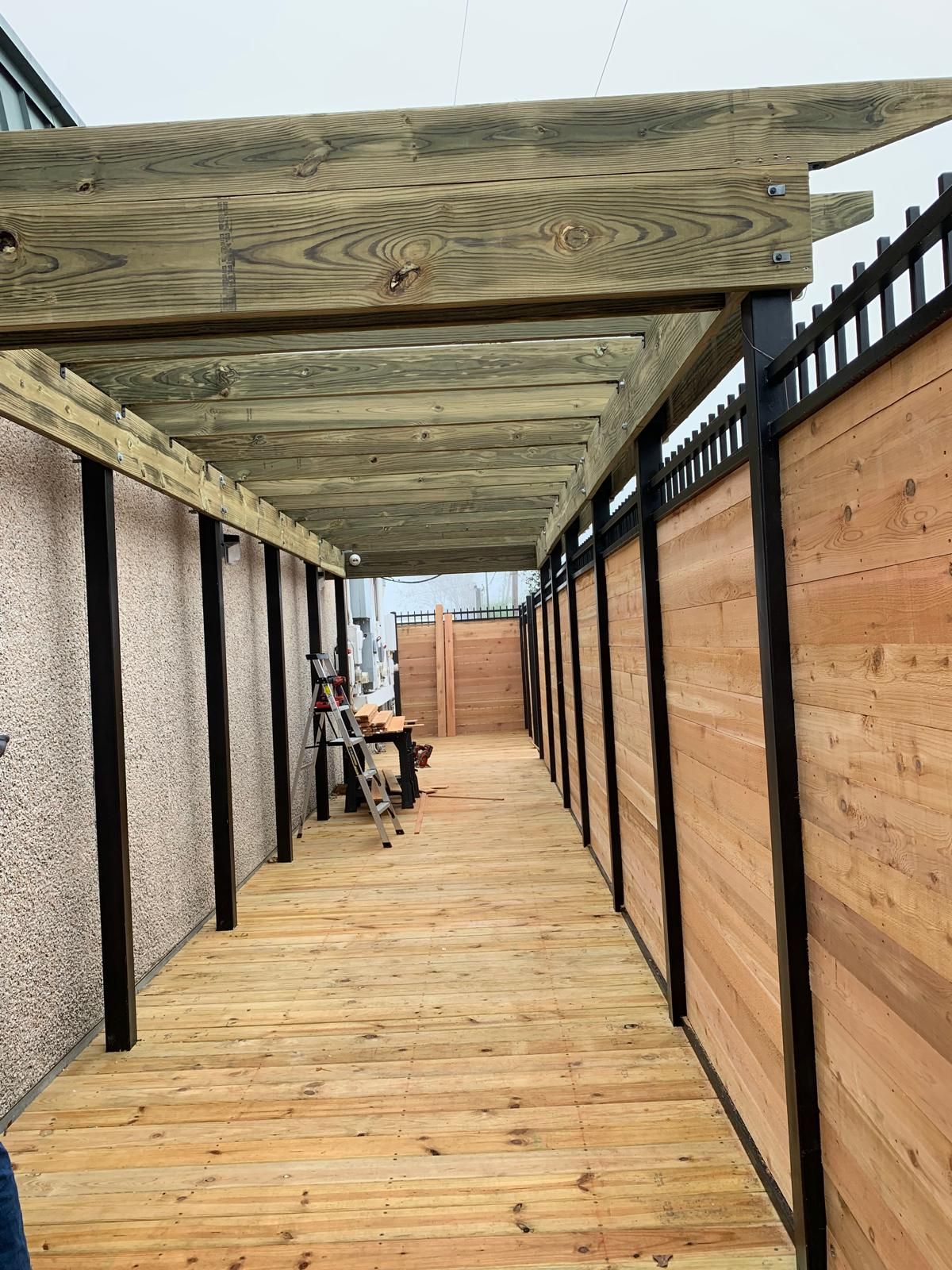 Wood-floored walkway with pergola overhead and wood/metal fences on both sides.