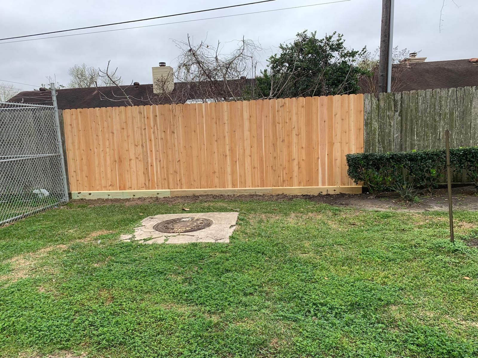 Backyard with wooden fence, chain-link fence, and grassy lawn. Manhole cover in foreground. Overcast sky.