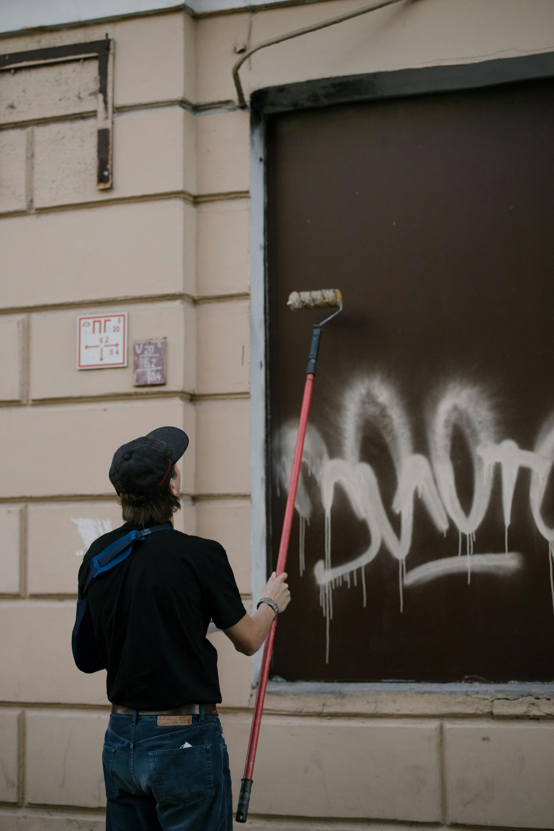 Person in black shirt and hat painting over white graffiti on a brown boarded-up window with a paint roller.