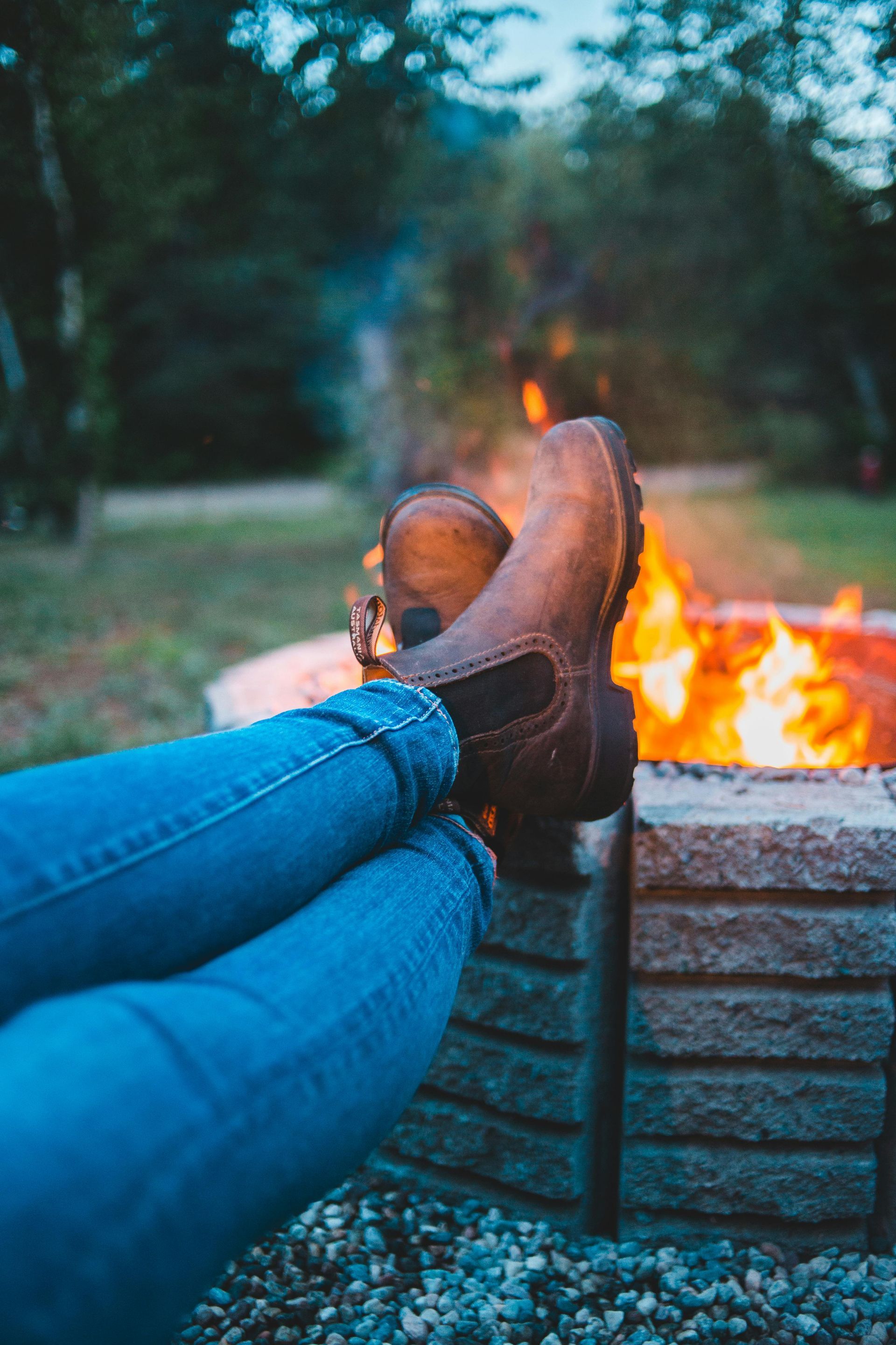 Person relaxing by a campfire, wearing blue jeans and brown boots.