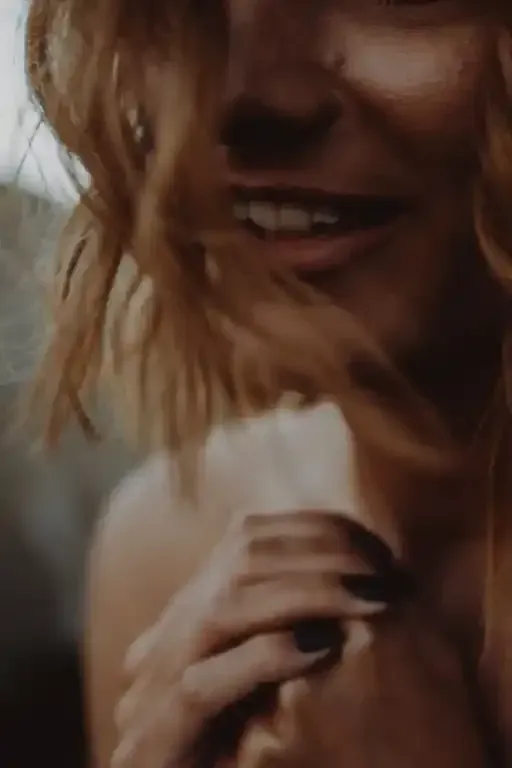 Donna sorridente, capelli mossi dal vento. Mani giunte, unghie scure. Parzialmente nascosta dai capelli.