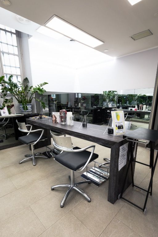 Interno del salone con postazione di lavoro in legno scuro e sedie girevoli. Piante e specchi sono visibili sullo sfondo.