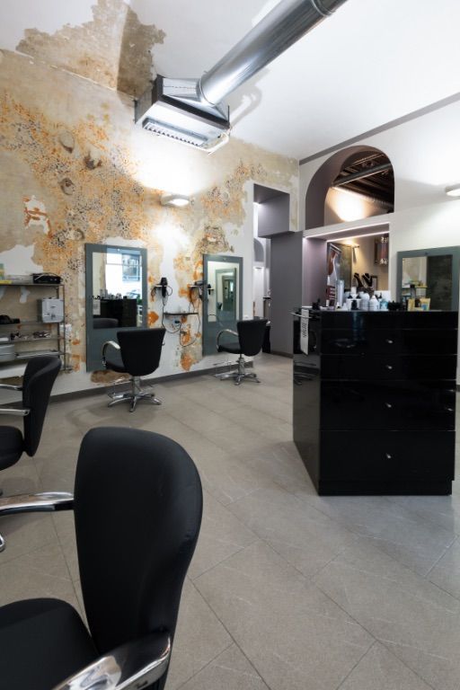 Interno di un salone da parrucchiere con poltrone nere, pareti rovinate dal tempo e illuminazione intensa.