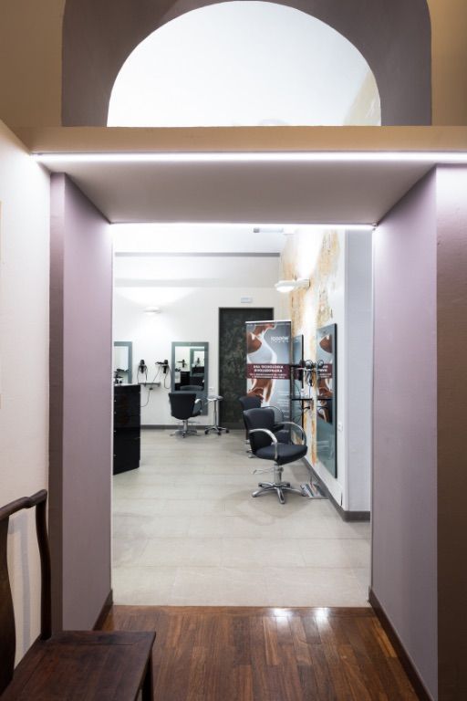 Corridoio che conduce all'interno di un salone da parrucchiere, con sedie, specchi ed espositori; luci soffuse.