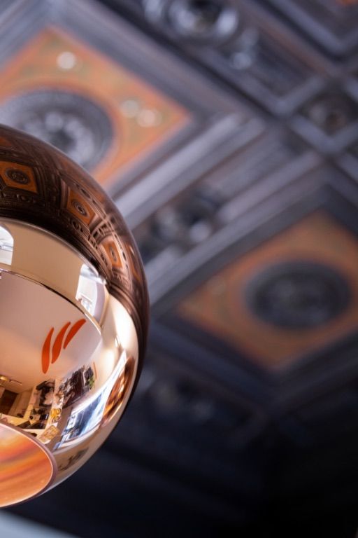 Una lampada a sospensione in rame riflette l'interno di una stanza. Sullo sfondo, un soffitto a cassettoni decorato.