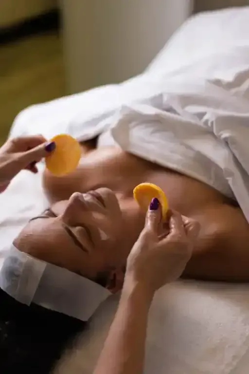 Persona che riceve un trattamento viso, sdraiata su un lettino da massaggio. Le mani tengono dei cuscinetti circolari arancioni vicino al viso.