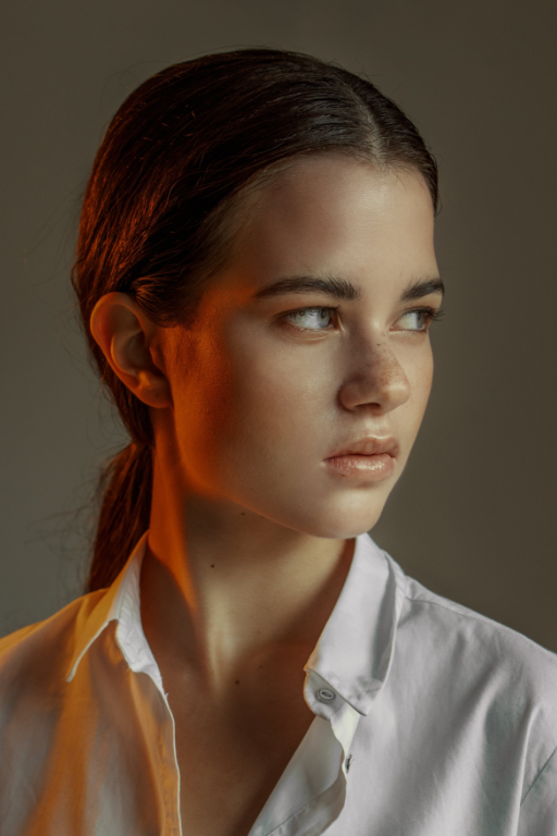 Donna con i capelli scuri tirati indietro, con una camicia bianca, che guarda di lato, illuminata da una luce arancione.