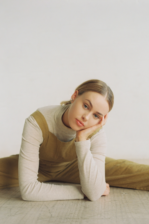 Una donna in tuta color senape è seduta sul pavimento con le gambe divaricate e la testa appoggiata su una mano. Sfondo bianco.