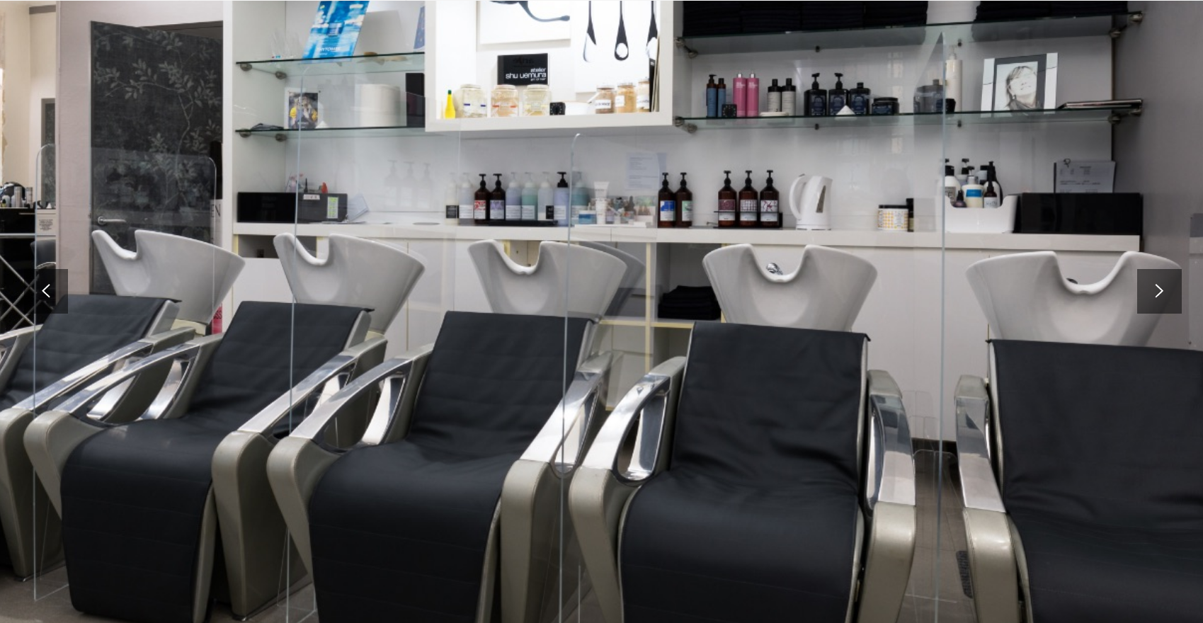 Interno di un salone da parrucchiere con postazioni di lavaggio e sedie disposte in fila. Decorazioni in argento e nero.