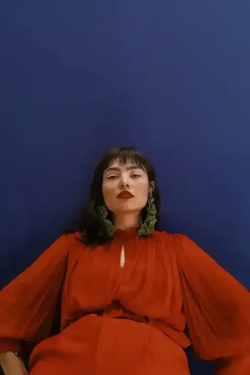 Donna con abito rosso e orecchini verdi su sfondo blu, che guarda la telecamera.