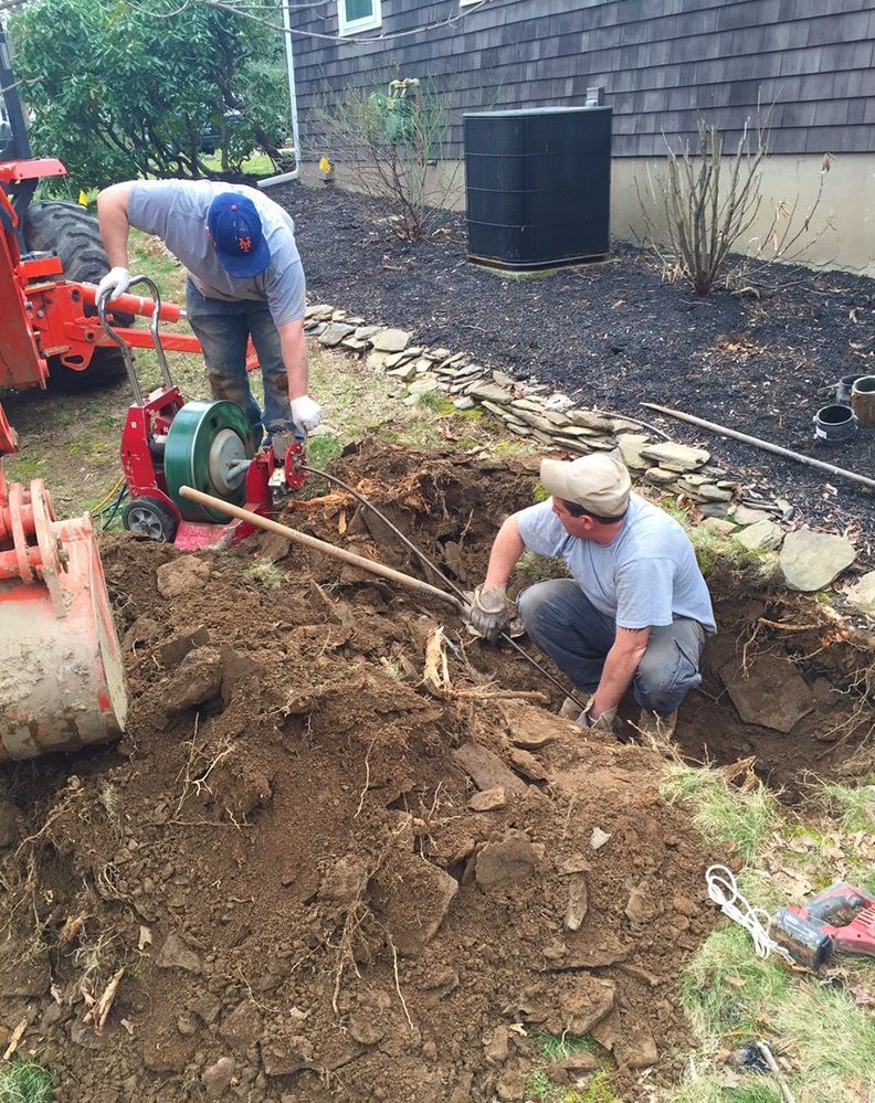 Two men are digging a hole in the dirt in front of a house.