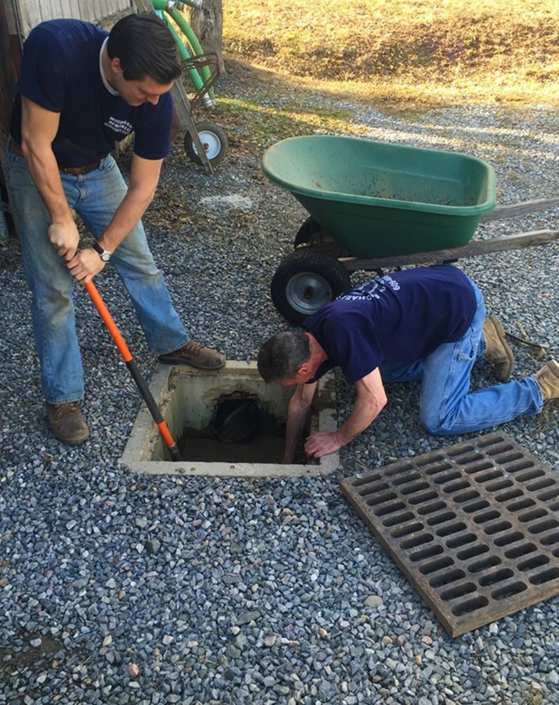 Two men are working on a drain with a wheelbarrow in the background.