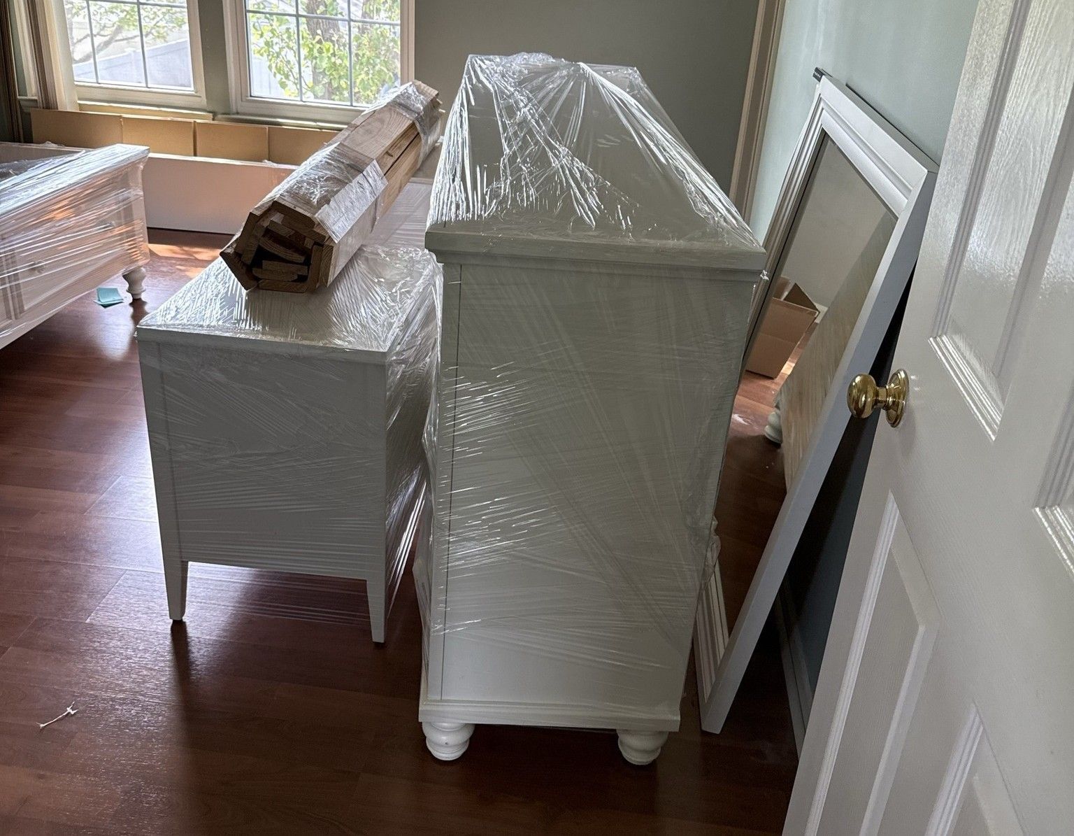 White furniture wrapped in plastic, in a room with hardwood floors. A large mirror leans against the wall.