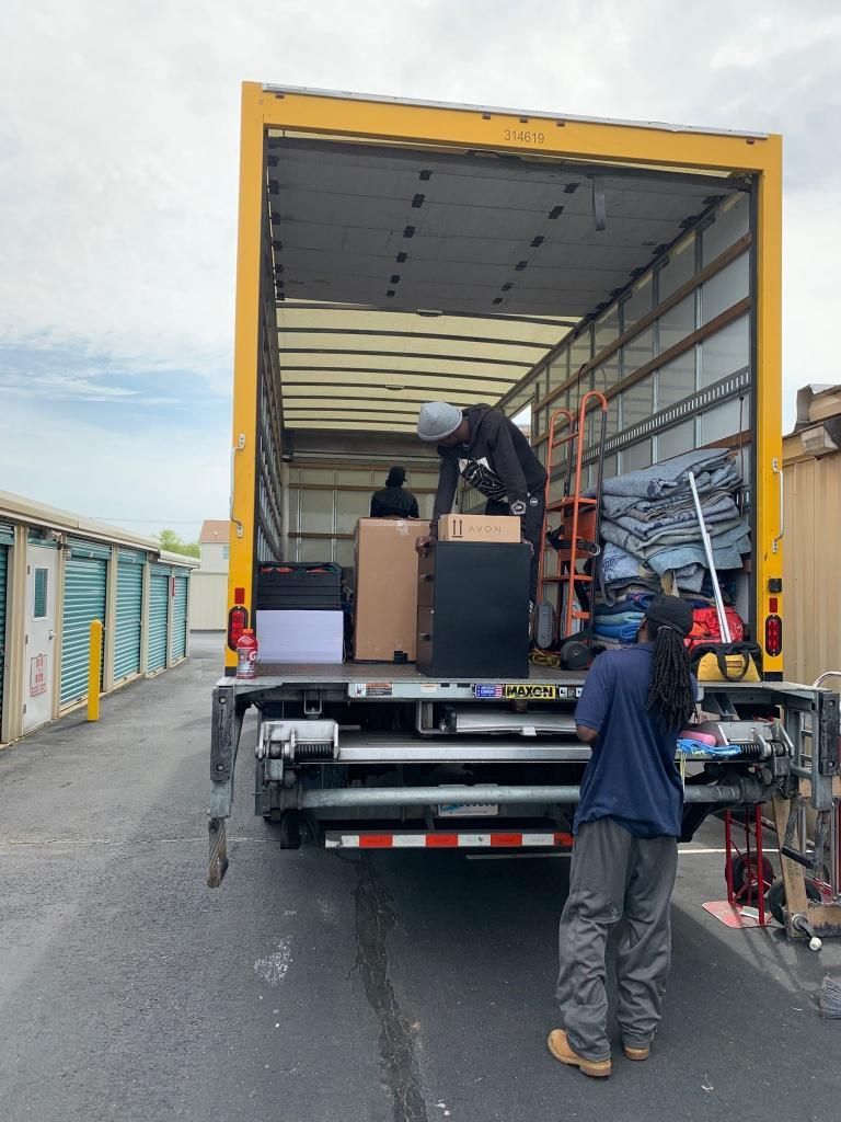 Movers loading boxes and furniture into a yellow moving truck. Exterior setting near storage units.