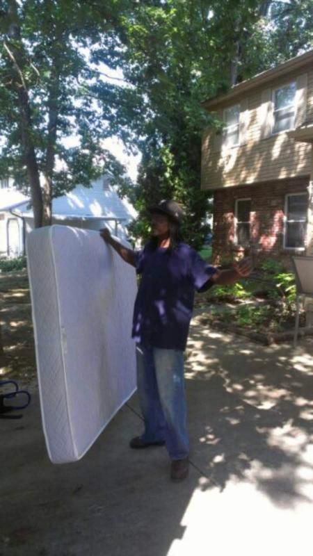 Person holding a mattress outdoors. Trees and a two-story house are in the background. Sunny day.