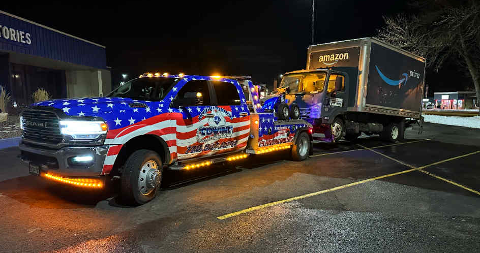 Tow truck with American flag design towing an Amazon truck at night.