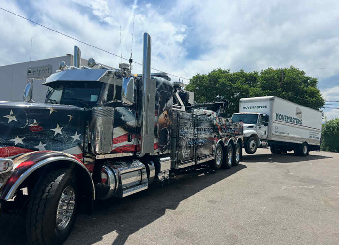 Patriotic tow truck towing a white delivery truck on a city street.