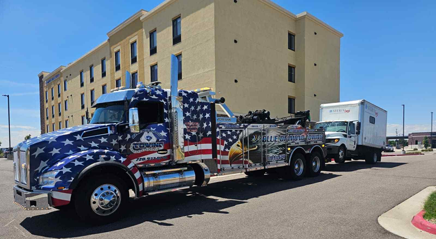 Patriotic tow truck towing a small white box truck in front of a beige hotel on a sunny day.