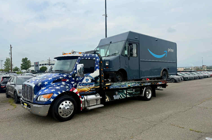A tow truck carrying an Amazon delivery van on its flatbed in a parking lot.