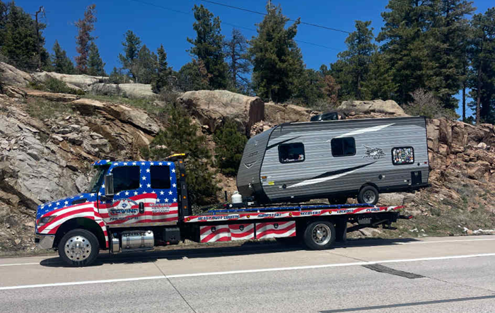 Tow truck with American flag design hauling a gray camper on a mountain road.