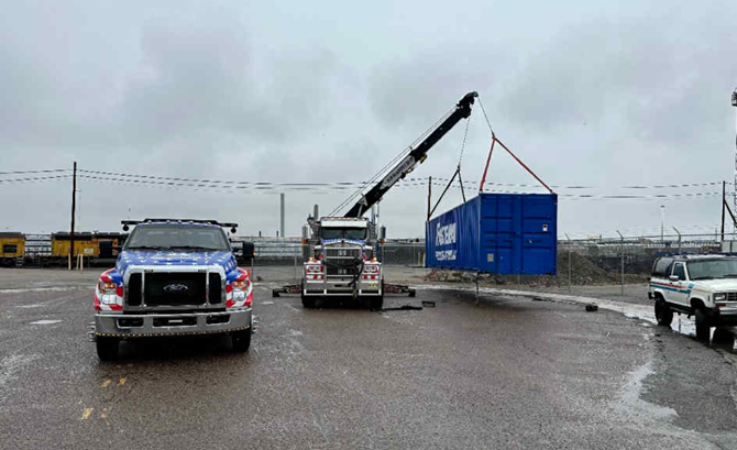A blue shipping container being lifted by a crane truck on a wet asphalt lot. Two other tow trucks nearby.
