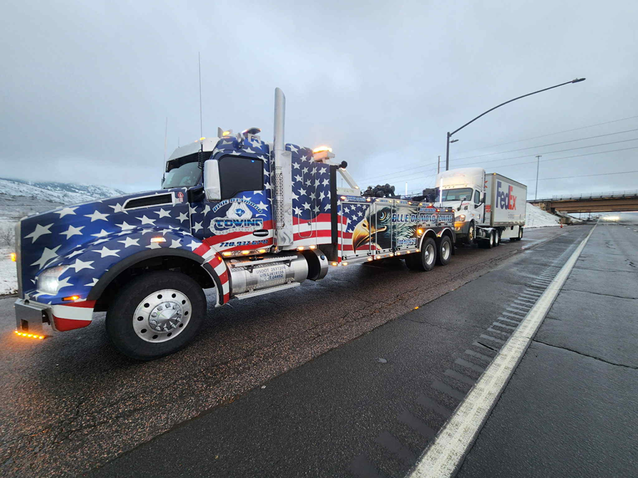 American flag-themed tow truck pulling a FedEx trailer on a highway, with snow in the background.