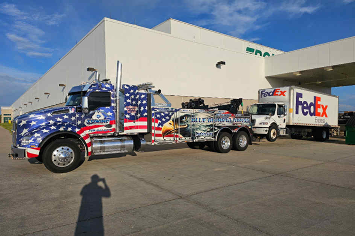 Tow truck with American flag design towing a FedEx truck outside a warehouse.