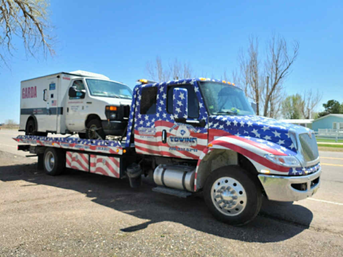 Tow truck with American flag design hauling a white armored truck down a sunny road.