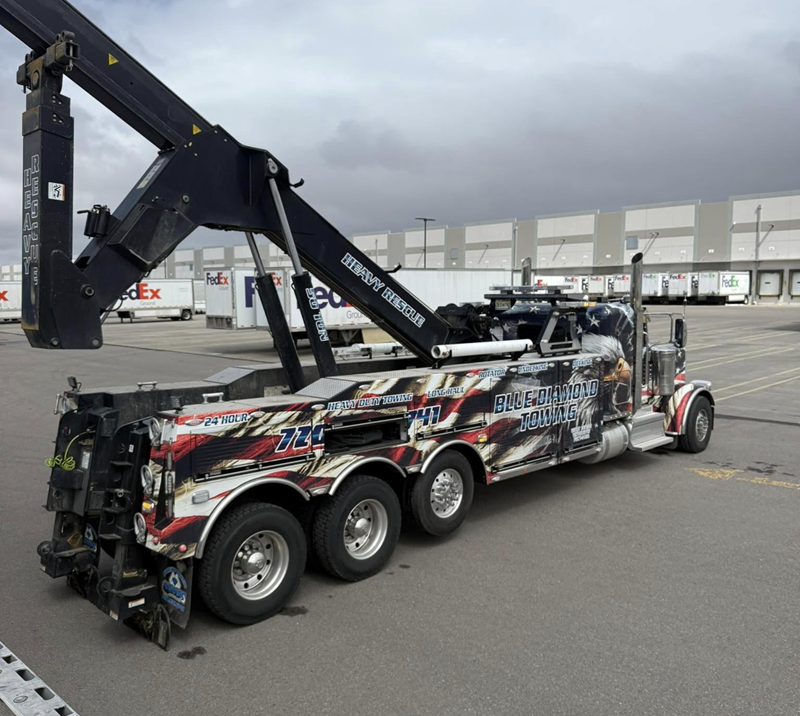 Tow truck with American flag design, parked in a lot near a warehouse, under a cloudy sky.