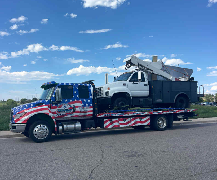 A patriotic-themed tow truck carries a white truck with a boom lift against a blue sky.
