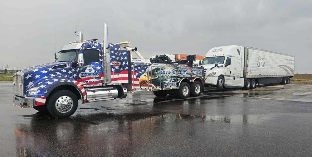 A patriotic tow truck pulling a white semi-truck and trailer on a wet, gray surface.
