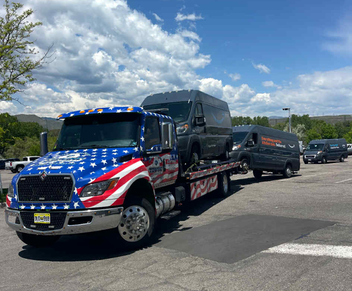 Tow truck with American flag design hauling two black vans in a parking lot on a sunny day.