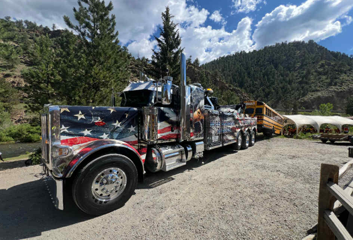 Tow truck with American flag design towing a school bus on a gravel road near a river and mountains.