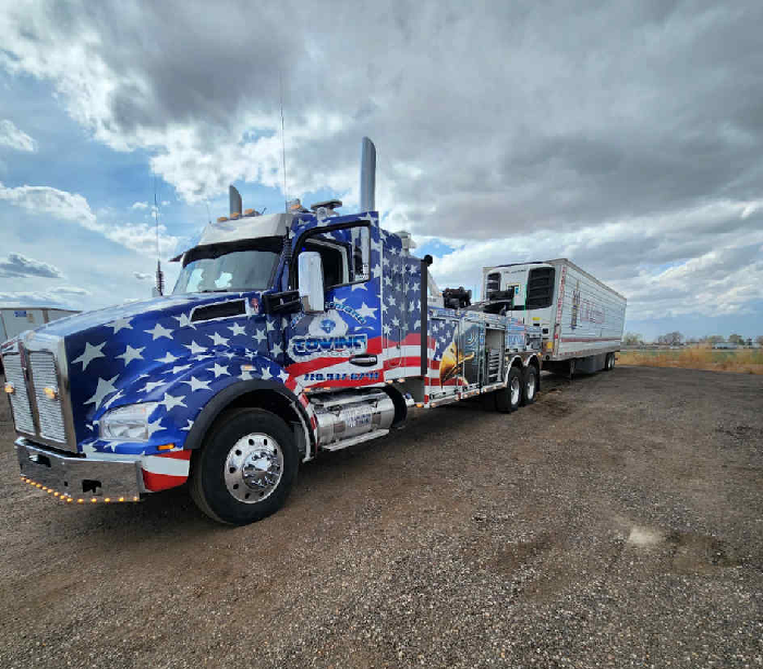 A patriotic tow truck hauling a trailer on a gravel lot under a cloudy sky.