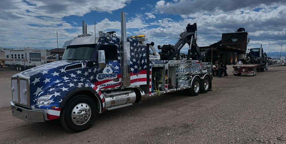 Patriotic tow truck on asphalt, towing another vehicle, under a cloudy sky.