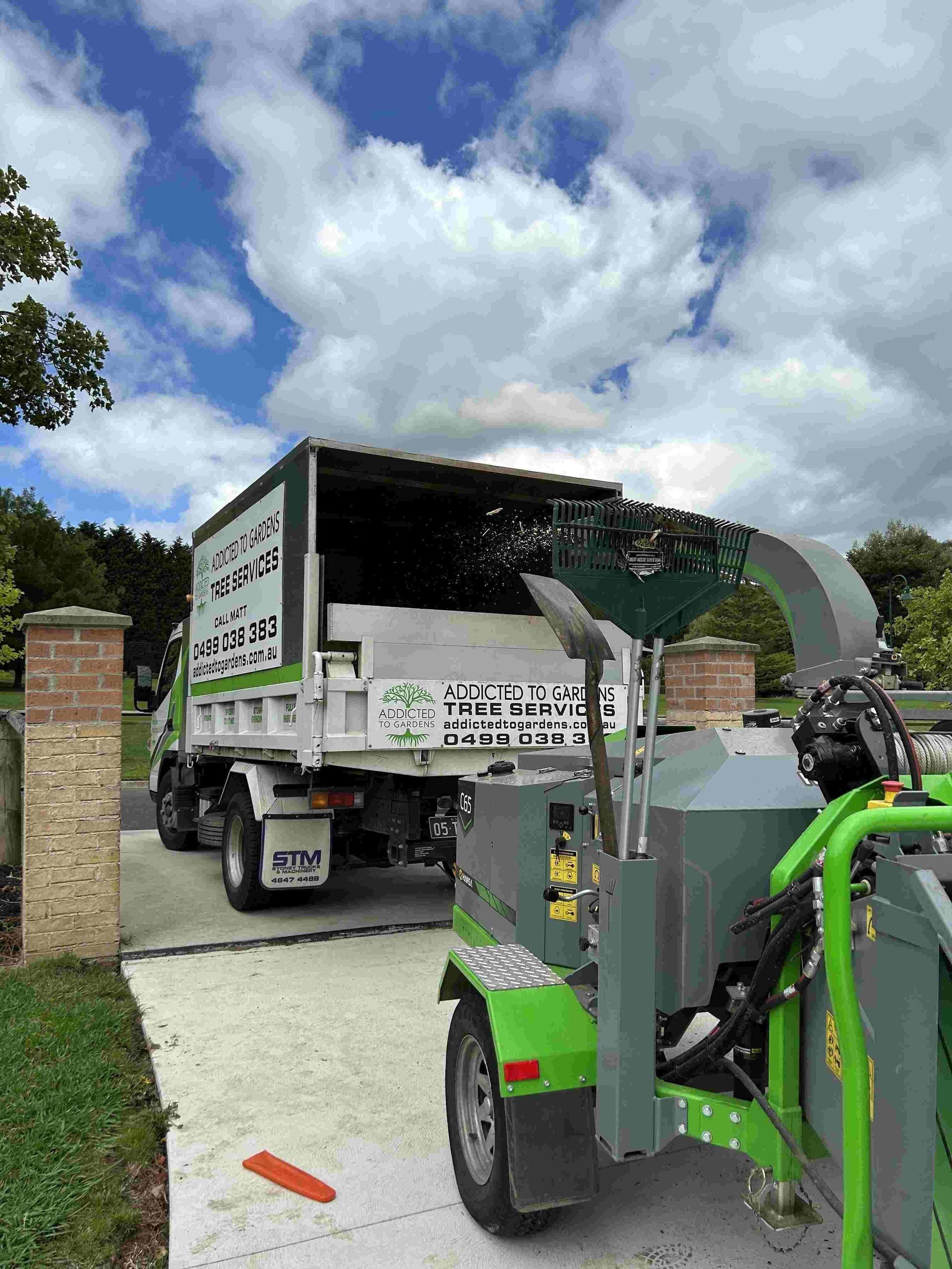 A tree service truck with a wood chipper.  The truck is unloading debris under a cloudy sky.